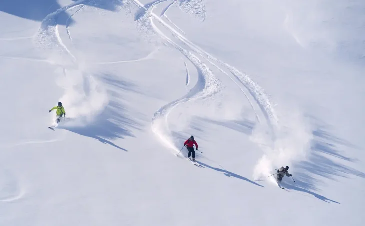 Three skiers photographed from above curve turn turns in fresh powder snow, leaving trails, on a ski holiday in Andorra