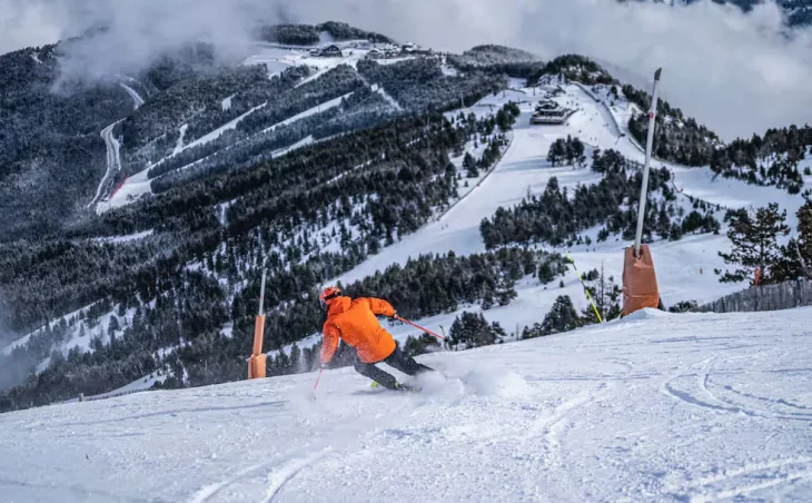 A skier dressed in bright orange skis down the slopes on ski holiday in La Massana, Andorra. Tree covered slopes can be seen in the distance.