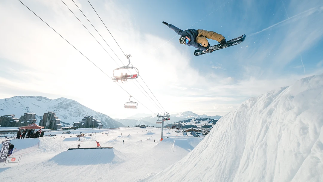 Snowboarder performing a method grab high in the Avoriaz snow park, with ski lifts and resort in the background.
